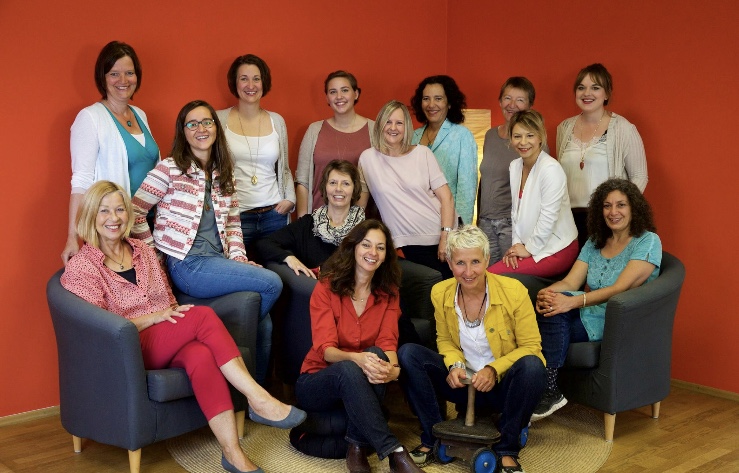 Das Bild ist in Herzform abgebildet. Es ist das Team des Frauen*notruf München darauf abgebildet. Es sind 14 Frauen abgebildet. Sie stehen und sitzen teilweise auf Sesseln.