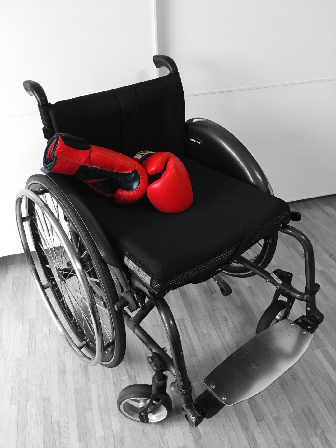 Auf dem Foto zu sehen ist ein schwarzer Rollstuhl. Darauf liegen zwei rote Boxhandschuhe.